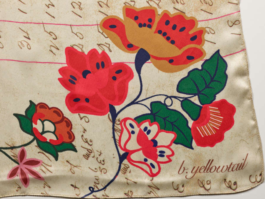 The scarf’s bottom right corner, showing a vibrant graphic rendering of red, green, and yellow flowers. The shiny cream surface has writing of numbers and the word “b. yellowtail”.