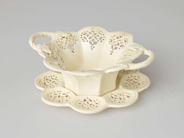 A cream-colored dish with two swirled handles and pierced decorations. Dish sits on a matching tray.