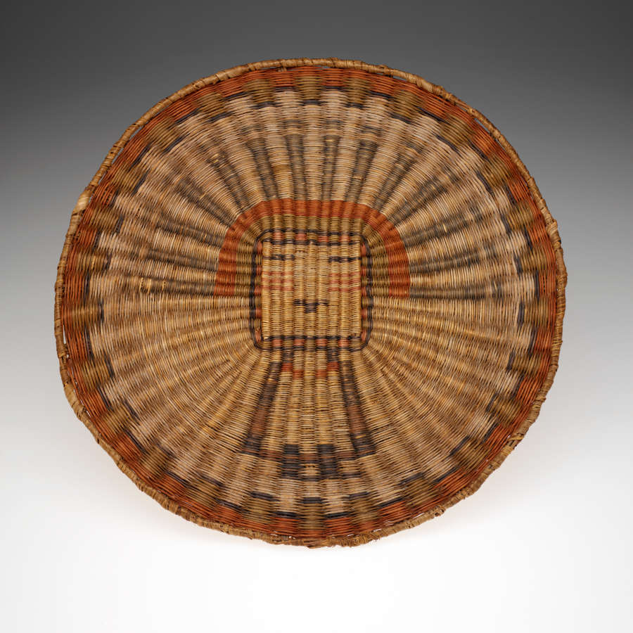 Circular basket, woven in a radial pattern with a bordered edge, in undyed, green, and red straw-like fibers. Woven in is an image of a necklaced head dressed human.