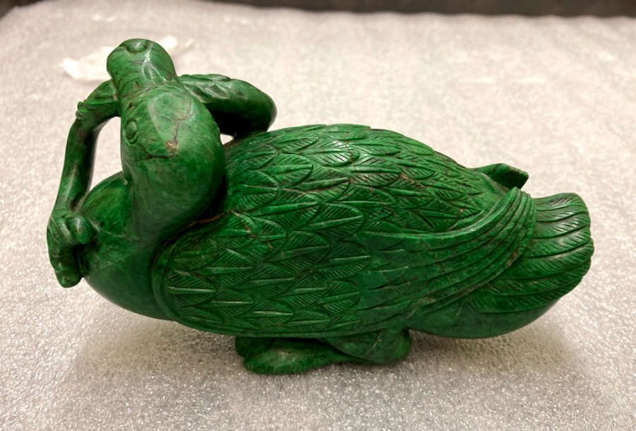 Green stone sculpture of a seated duck, its legs tucked under its body and its mouth holding a fish, looking away from the viewer. The duck’s feathers resemble leaves. 
