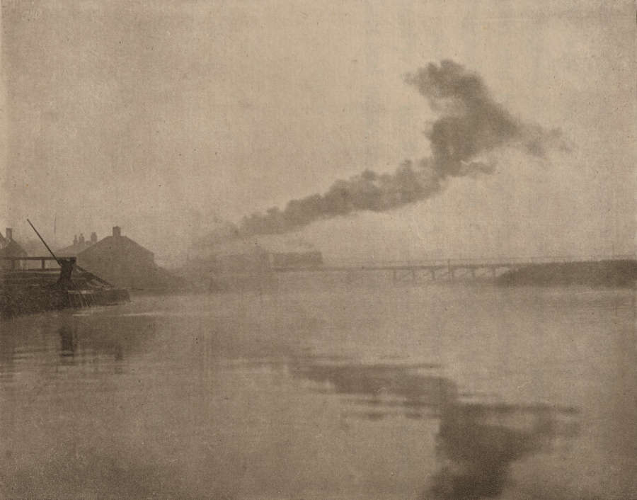 A black and white image of a house to the left of a calm body of water with a cloud of smoke coming from the distant bridge.