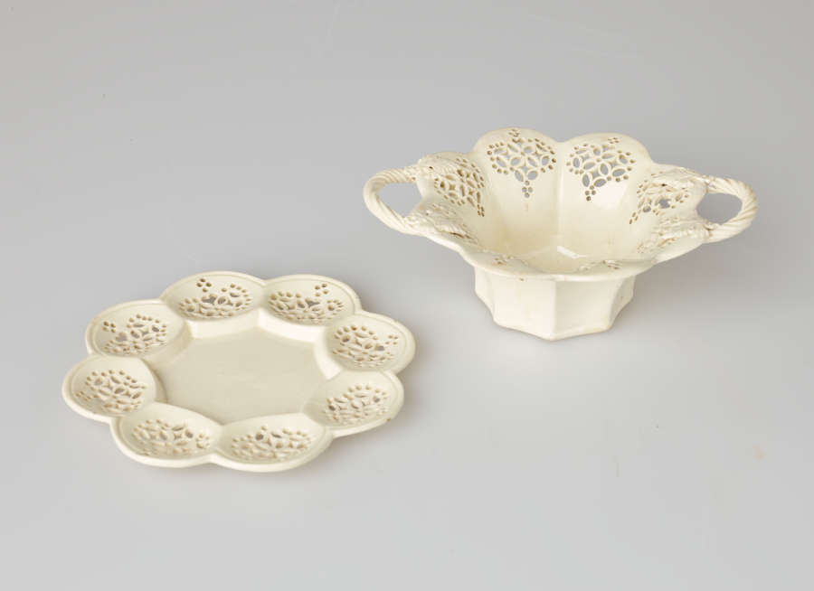 A cream-colored dish with two swirled handles and pierced decorations. Dish sits on a matching tray.