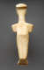 A smooth, cream colored figurine viewed from the back, depicting a flattened head, broad shoulders, and a slender lower body. 