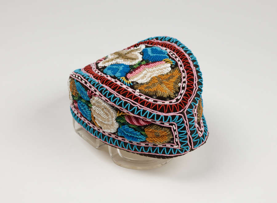 An almond shaped embroidered cap, which features 3 blocks of blue, white, green, and pink floral embroidery which are framed and divided by blue, pink, and white geometric patterns.