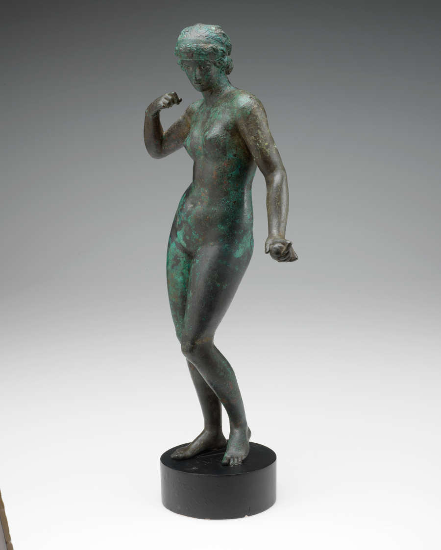 Quarter-view of a tarnished bronze statue of a standing crowned nude woman with one arm bent towards her shoulder and the other held out, standing on a circular pedestal.