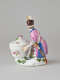 A sculptural figure kneeling next to a basket. Figure wears bright, elaborate clothing with a hat with a feather. There are floral decorations on the figure’s clothing and basket.
