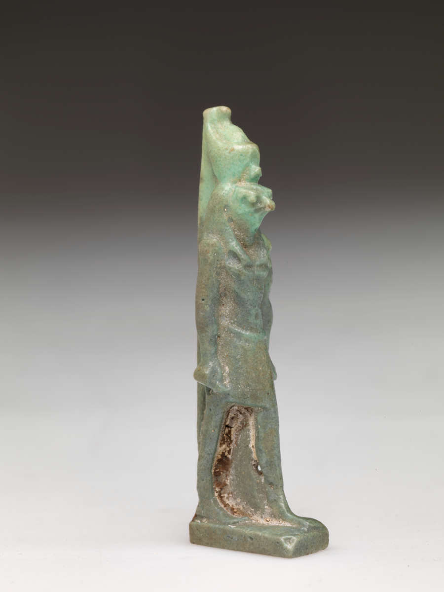 Partial side-view of light-jade sculpture of a standing person with a tall head, narrow torso, arms straight. The left leg has brown specks on it while the headdress is pale.