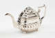 A silver teapot with a decorative angular handle, a rounded square body and spout with sculptural decorations.