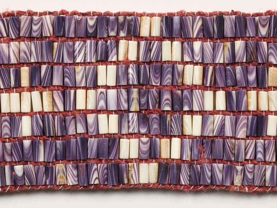 Detail-view of the patterned beaded belt constituted by rows of uniformly sized cylindrical beads carved from shells with purple and white striations, woven together with maroon thread.