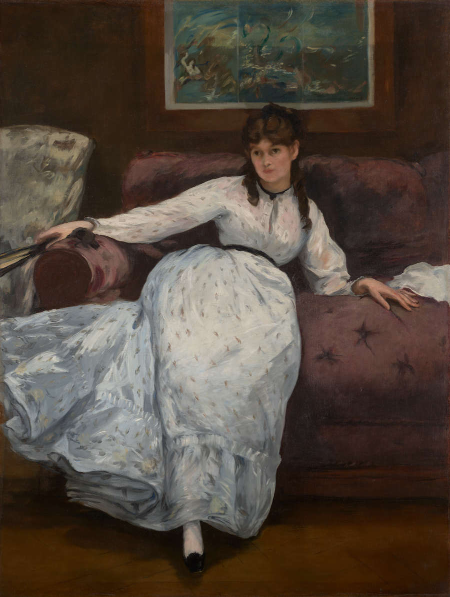A woman with dark hair and a calm expression reclines in a long white dress on a red sofa, one arm outstretched holding a fan, beneath a framed blue turbulent seascape painting.