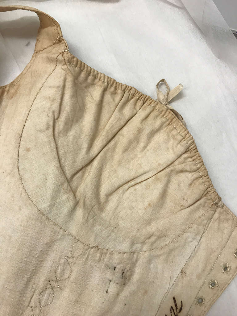 Close-up of slightly discolored cream fabric garment with delicate stitching, gathered neckline with small looped ribbon and small eyelets on the side. 