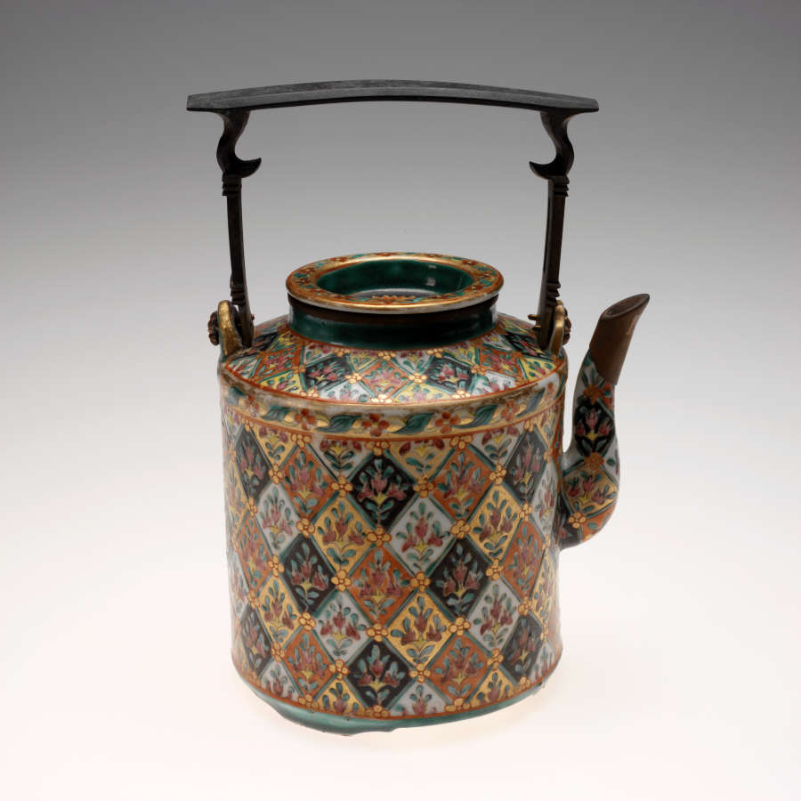 A cylindrical teapot with an elaborate design of flowers inside diamond shapes of varying colors. A thin, square shaped handle with a flat top sits above the teapot.
