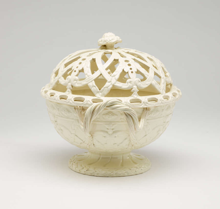 A cream-colored basket with sculptural woven handles, sculpted foot, and a lid with cutout designs.