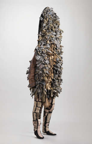 Side view of full-body sculptural garment in earthy tones engulfing the upper body in dense glove or finger-like forms with a sweater sleeve hanging out and legs clad in patchwork.