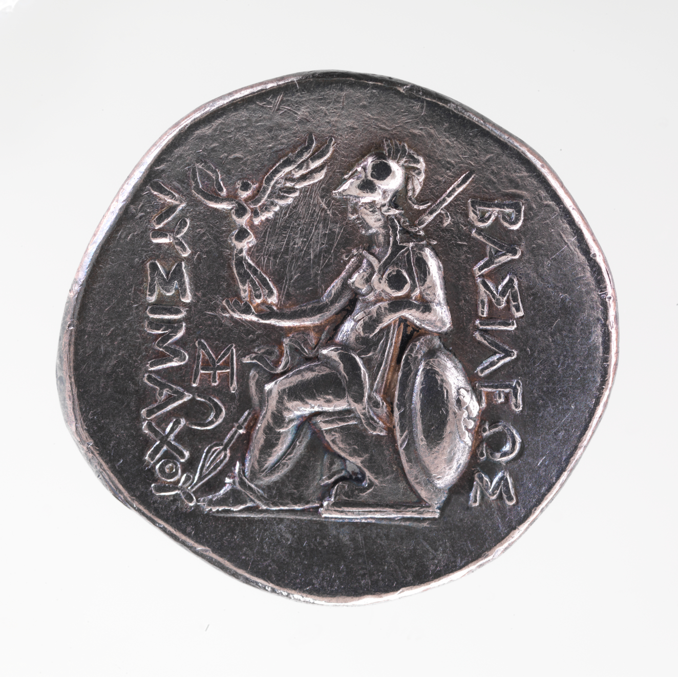 A silver coin featuring a central engraving of a seated figure wearing a helmet, propped on a shield, holding a small winged figure, with Greek inscriptions framing the design.