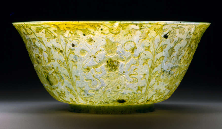 Jade bowl with sloped edges and a subtly flared mouth, with ornate floral etchings and a figure photographed in lighting making its surface appear bright white and yellow. 