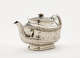 A silver teapot with a decorative handle, a rounded square body and spout with sculptural decorations.