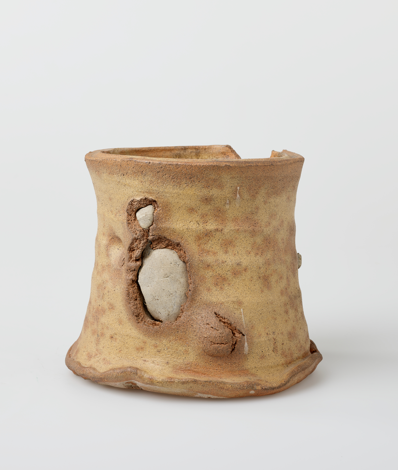 Cylindrical beige vessel with a lightly weathered surface and an uneven rim featuring a prominent chip. Two irregular oval openings expose smooth, pale gray embedded fragments beneath the surface.