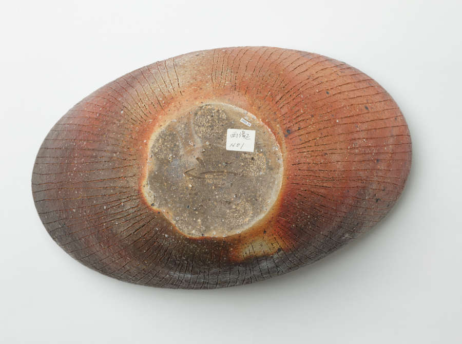 Bottom of an oblong stone bowl with shell-like etchings. Its body is dark orange-red, fading into darker green-grays. The circular, gray base has a white rectangular sticker.