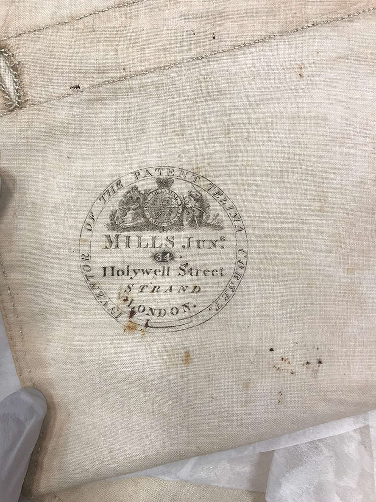 A close-up of an old textile with faded black ink stamp in a circular design and text reading “Inventor of the Patent Telima Corset. Mills Junr, Holywell Street, Strand, London.”