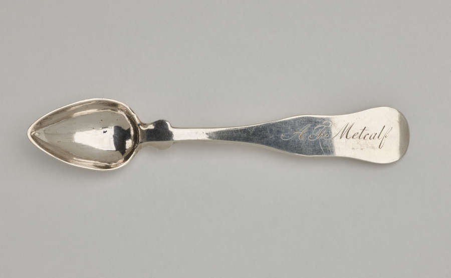 A silver salt spoon the bowl comes to a pointed tip, the neck is thin, and the handle is thicker and rounded at the end.
