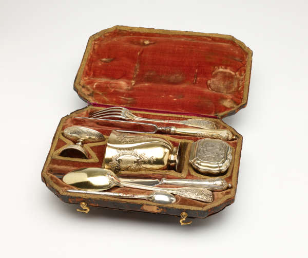 A traveling set with silverware. The traveling case has an orange colored fabric. There are multiple indents holding the silver, steel, and gilded cutlery and vessels.