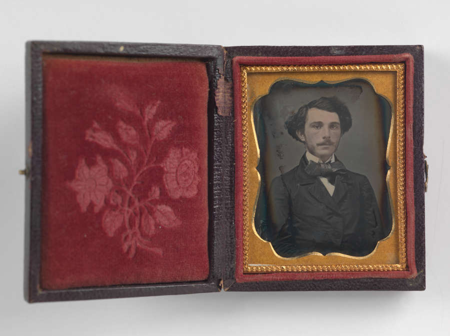 An open black case with a rose imprint in red velvet on the left side, and an orange framed black and white portrait of a man in a suit and bowtie on the right.