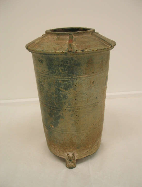 Side-view of a tall and narrow green-gray cylindrical terracotta urn with a narrow mouth and wide, upturned saucer-like neck, tapering into a narrow base, with one ornately carved foot visible. 