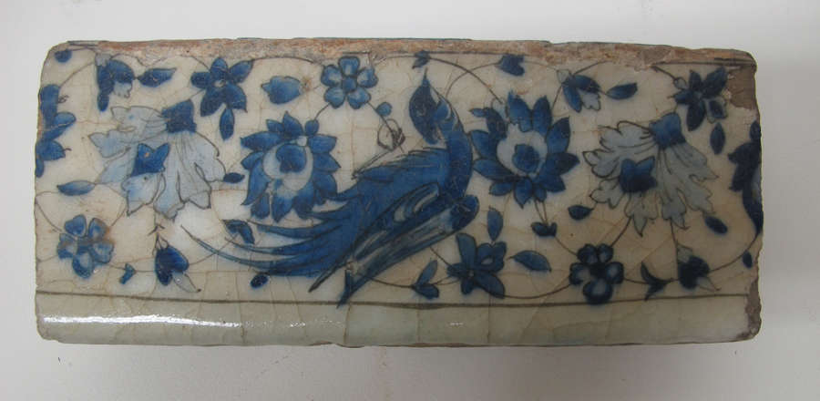 A horizontally long rectangular tile fragment. The top is worn, brown and the tile is an off-white color with indigo designs of a bird against a symmetrically arranged floral pattern. 