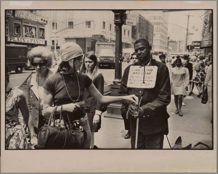 A grayscale photograph of a light-skinned person putting change into the cup of a dark-skinned person, who wears a sign around their neck that reads “I AM BLIND AND DEAF.”