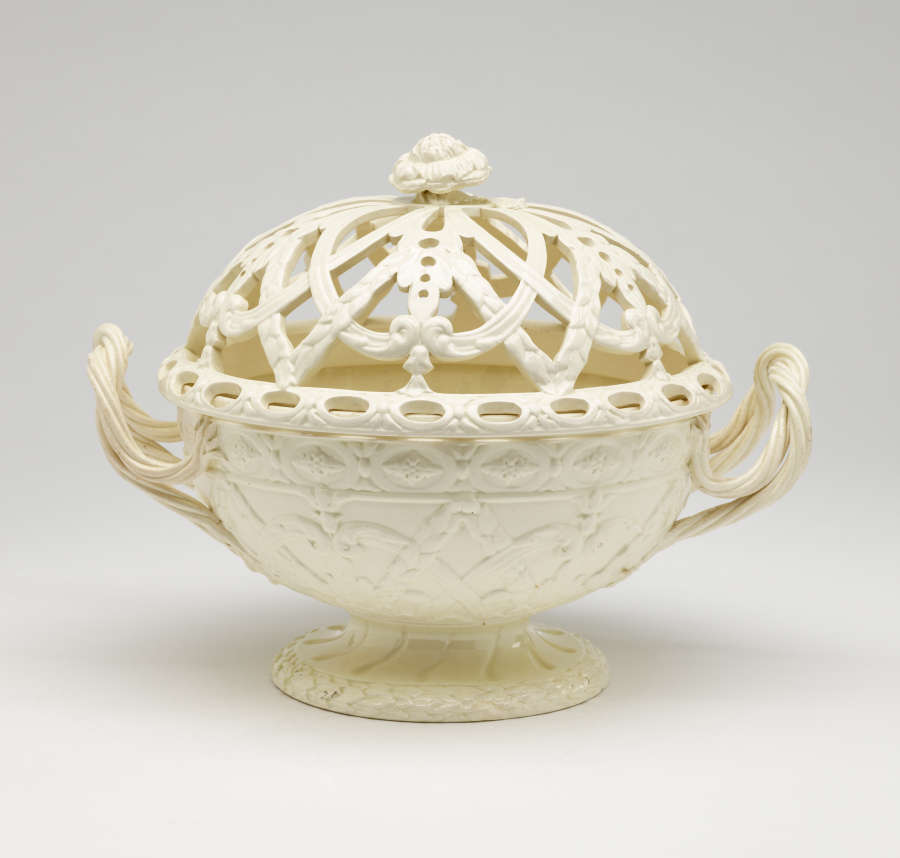 A cream-colored basket with sculptural woven handles, sculpted foot, and a lid with cutout designs.