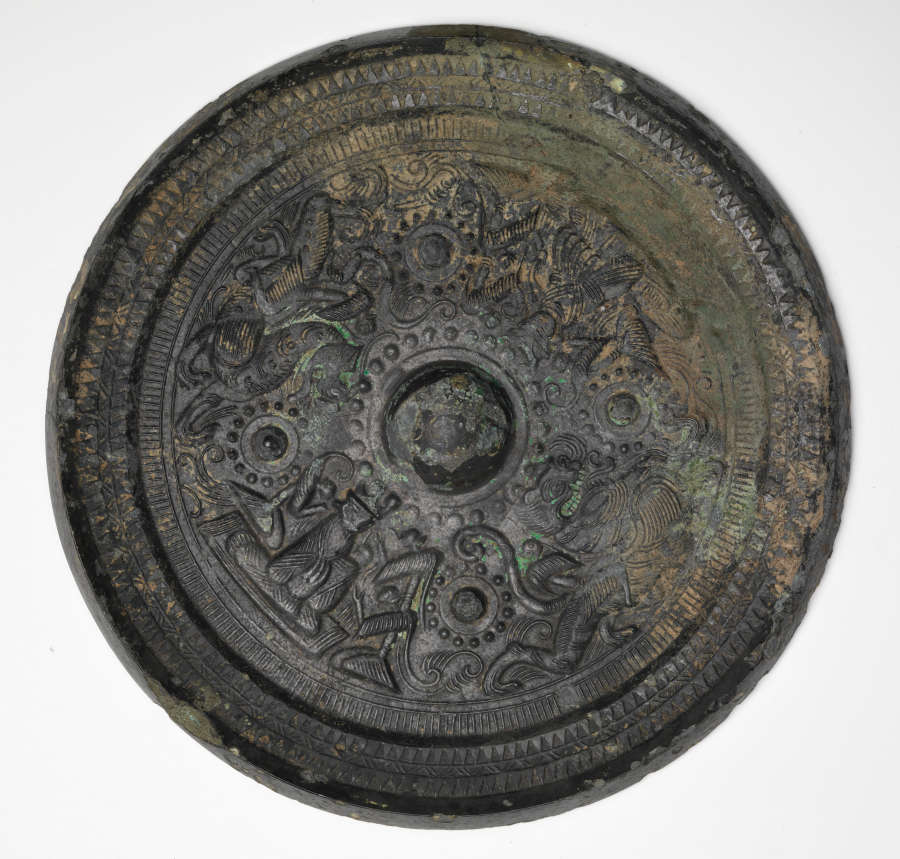 Black, round and corroded back of a handless mirror, embellished with a thick raised border, concentric illustrations and a domed center. It also has a greenish-yellow patina.
