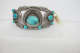 Silver and turquoise bracelet on a rectangular white base. The bracelet features two rows or ornately patterned silver connected by turquoise stones enveloped by silver crescents. A yellow label reads “67.170”.