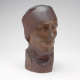 Three quarter front view of a dull carved wood sculpture depicting a serene face wearing a cloth around its head and neck. The bust features smooth contours and a polished finish.