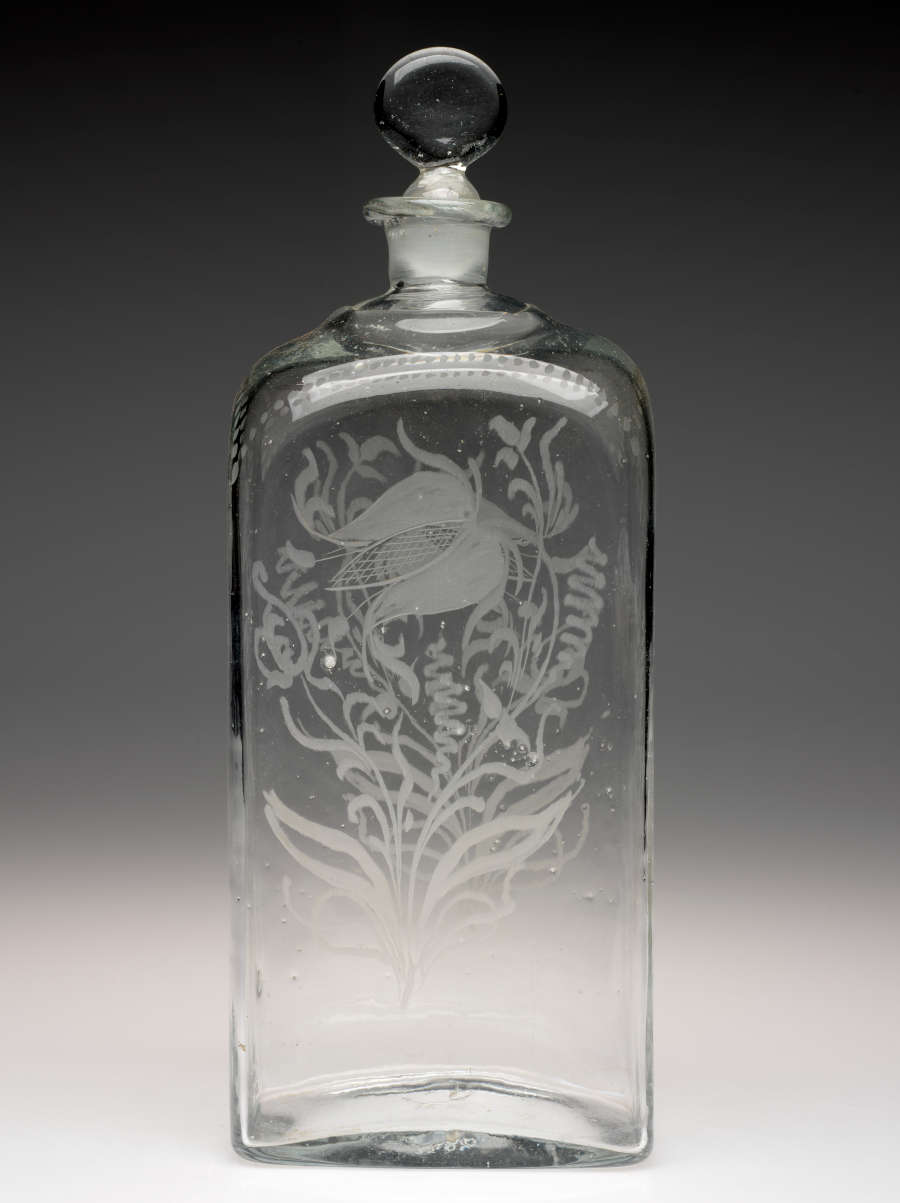 Tall rectangular clear bottle with rounded stopper. A floral design with leaves and swirling lines is etched on the front and back.
