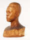 Three quarter front view of a glossy, carved wooden bust depicting the head of a male figure with afrocentric facial features. He has a stern expression and an intense gaze.