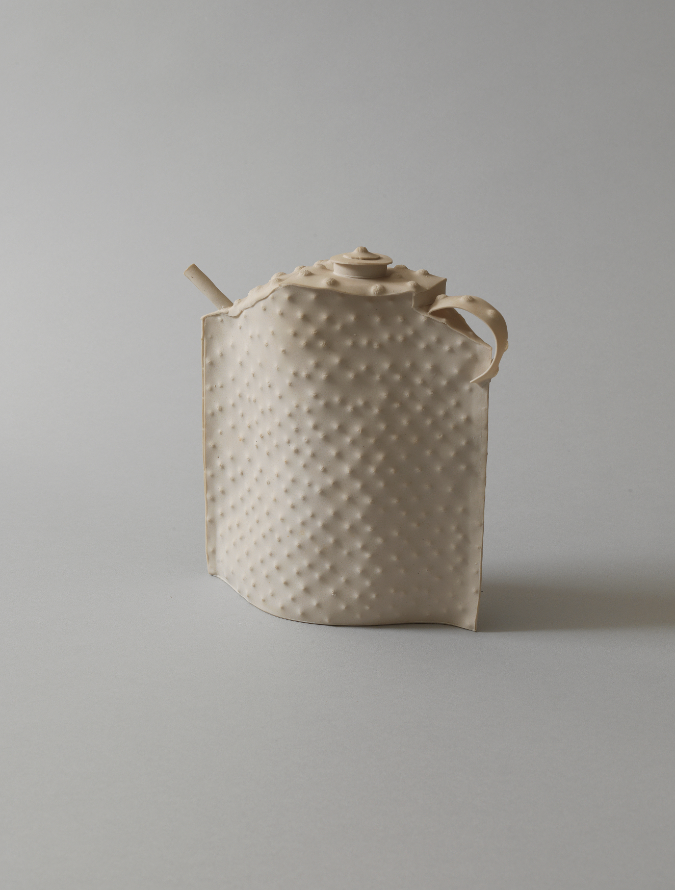 Minimalistic teapot with a dimpled surface, slightly flattened form, narrow spout, small curved handle, and a small coiled lid, all made in smooth, cream-colored matte ceramic.