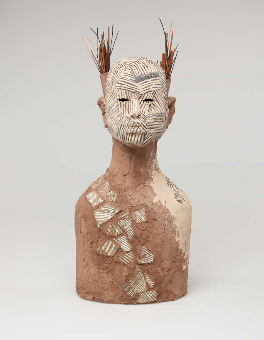 Brown clay bust of a figure with a grass and stick headdress. Diagonal lines of patterned patches of offwhite run across their chest and cover their face.