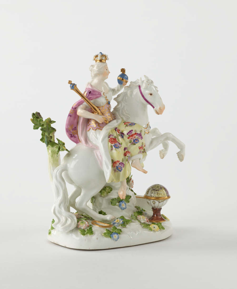 A sculptural figure on a rearing horse in historical clothing that is yellow, pink, and white with floral decorations. The figure is holding a staff and ball.
