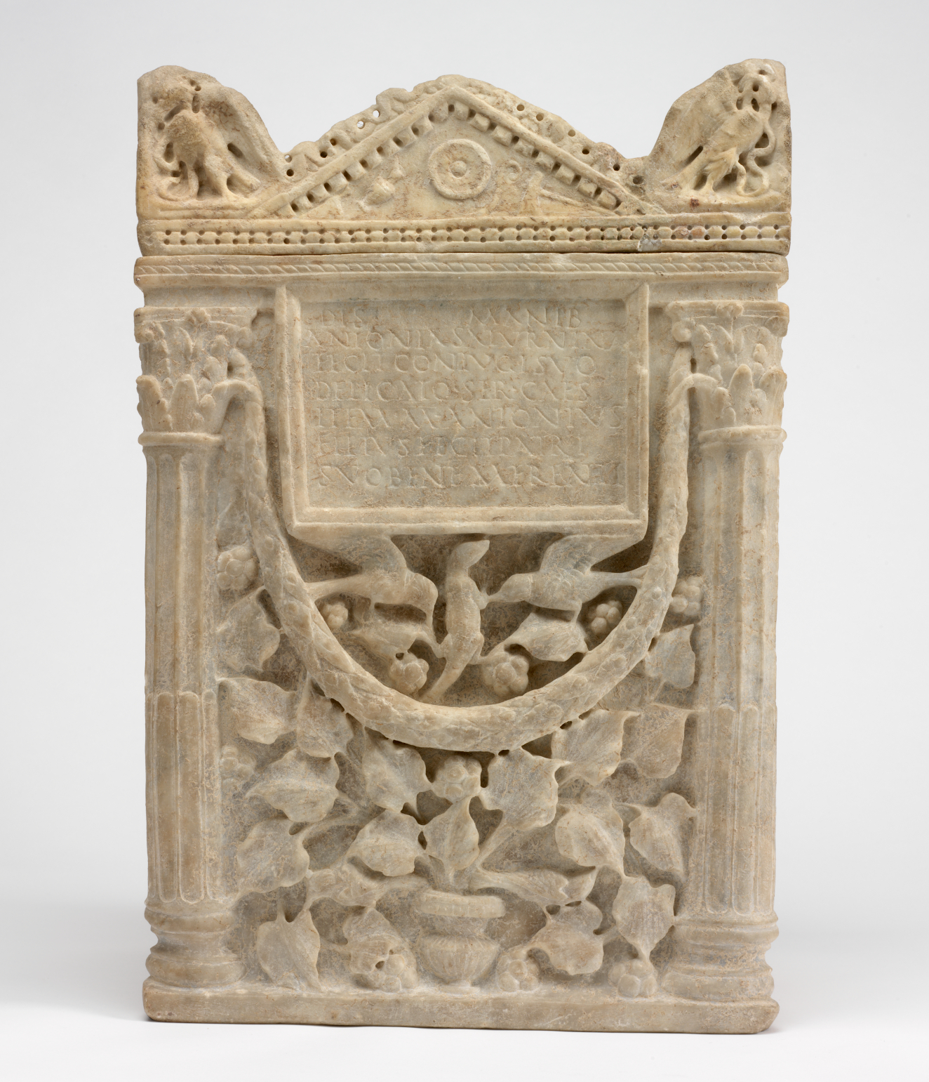 A rectangular carved stone monument featuring a triangular sloped roof supported by columns flanking a carved inscription above intricate, dense leafy vine and animal motifs emerging from the bottom. 