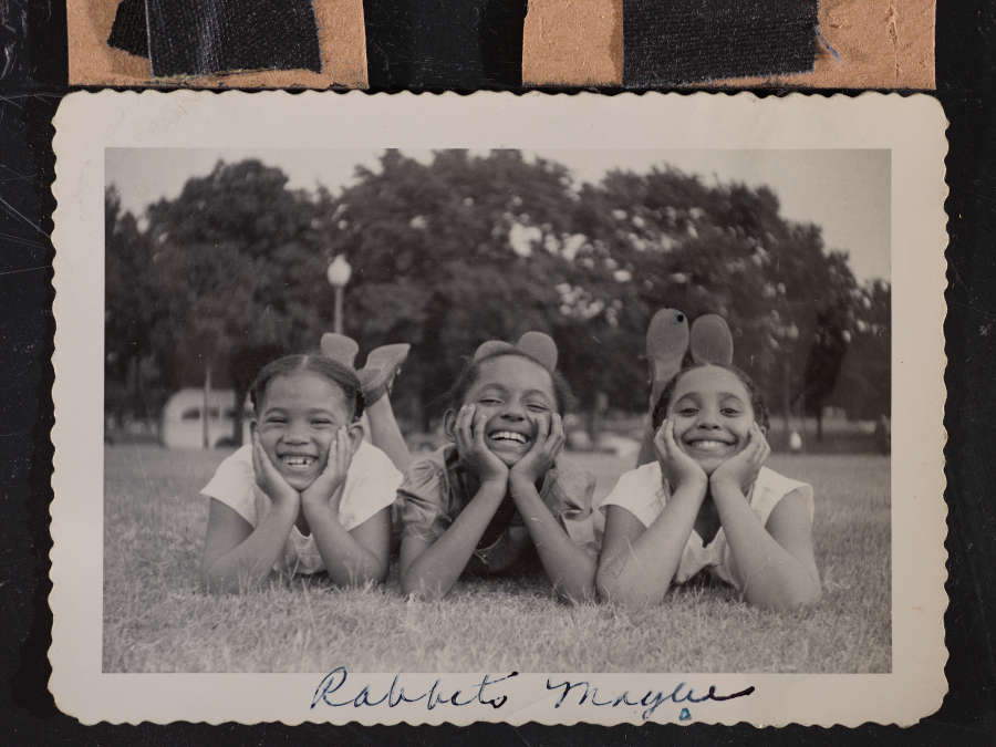 Black and white snapshot of three medium-skin-toned girls, smiling and lying on their stomachs in the grass, hands supporting their heads. There are trees and cars in the distance.