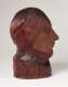 Profile view of a vibrant, carved wood sculpture depicting a face wearing a draped cloth around its head and neck. The bust features smooth contours and a polished finish.