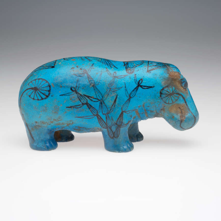 A side view of a blue sculpture of a hippopotamus with black illustrations of grasses and aquatic plants painted on it. Some chipping reveals its brown unglazed surface.