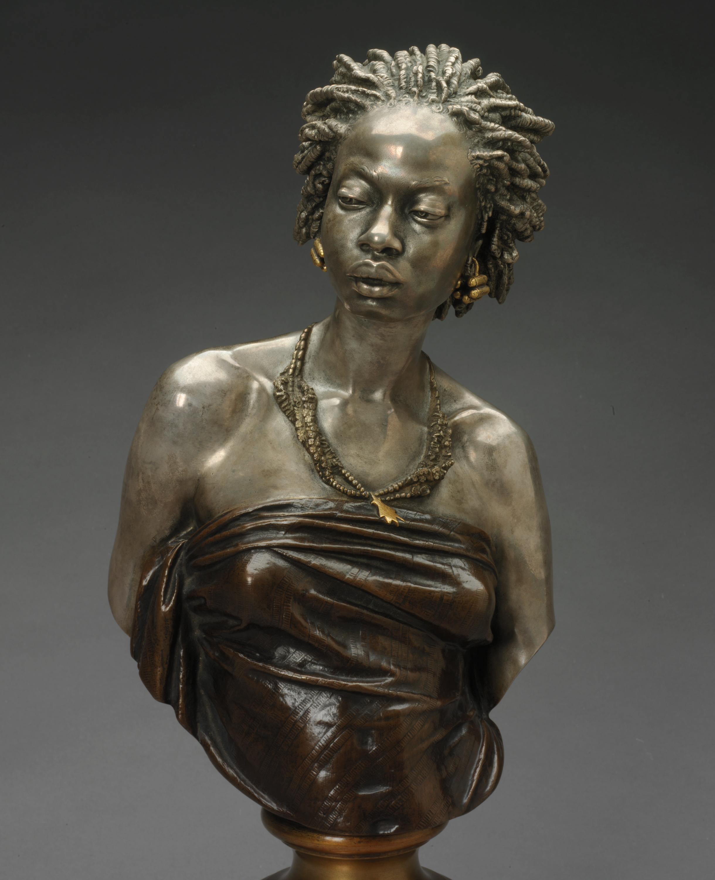 Muted bronze bust of a woman gazing downwards with hair in twists, gold earrings, and beaded necklaces accentuating her collarbones above a dark strapless tightly-wrapped top mounted on a gold base.