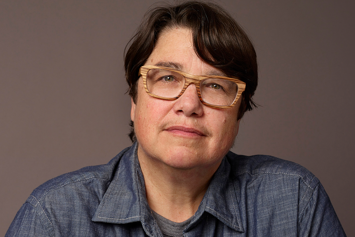 Headshot of artist Catherine Opie, with short hair, glasses, and a collared denim shirt, looking directly at the camera.