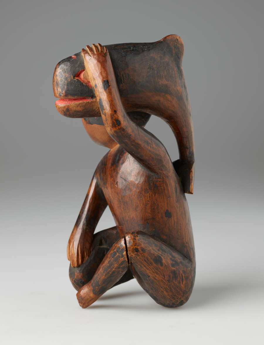 Wooden sculpture of a kneeling bearded man looking aside, holding a small whale against his head. Both man and fish are detailed in partially worn red and black paint.