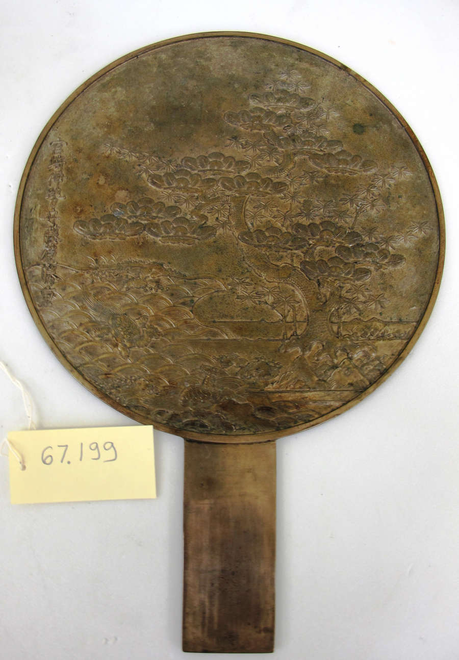 The reverse of a round bronze hand mirror with a worn surface engraved with trees and mountains, with a paper tag labeled "67.199".