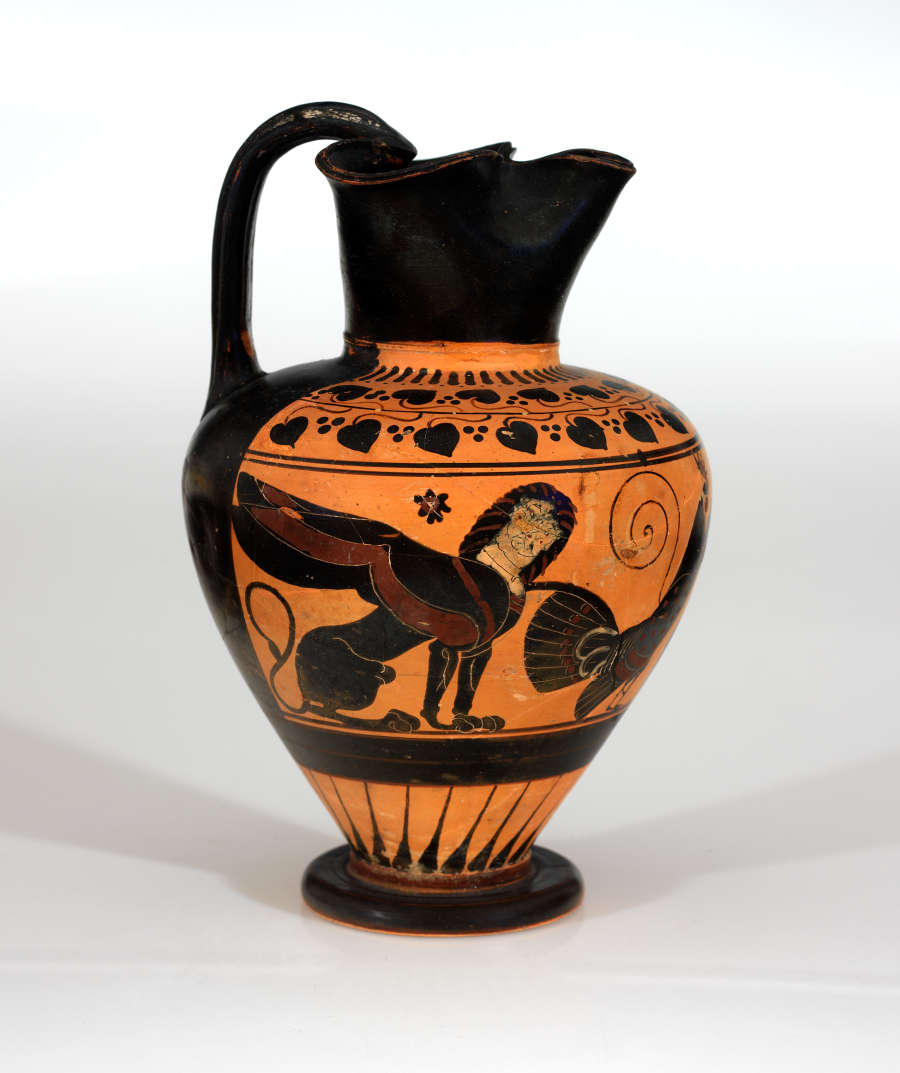 Tall black and orange onion-shaped jar with a pinched mouth forming a spout and a handle. It is decorated with natural and geometric patterns and illustrations of a sphynx.