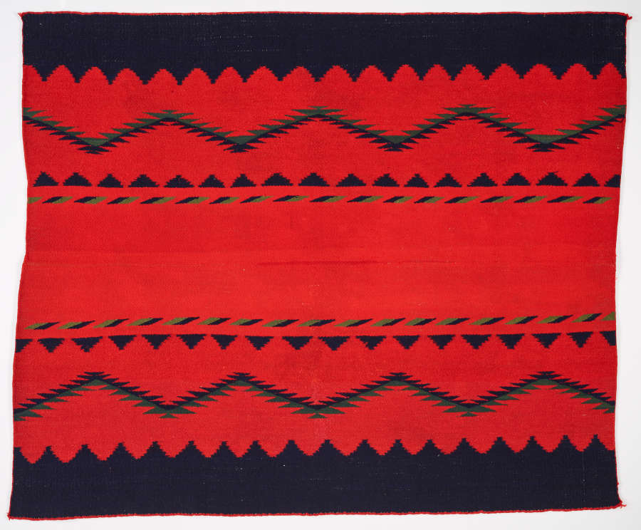 Red woven tapestry with blue-black geometric triangles and zigzag patterns. The tapestry has a blue-black jagged trim and the patterns are horizontally mirrored along the center.