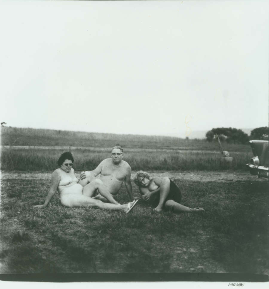 Black and white photo of an awkward, naked, light-skinned family reclined on grass. The lumpy woman only wears sandals. The equally lumpy man appears naked. The adolescent child wears shorts.
